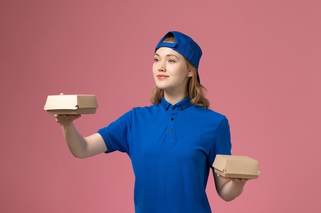 Weiblicher Kurier der Vorderansicht in der blauen Uniform und im Umhang, die kleine Liefernahrungsmittelpakete an der rosa Wand halten, Dienstjobuniformdienstmitarbeiterarbeiter