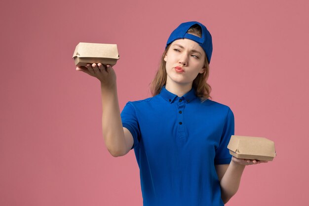Weiblicher Kurier der Vorderansicht in der blauen Uniform und im Umhang, die kleine Liefernahrungsmittelpakete auf rosa Wand, Dienstuniform-Dienstleistungsunternehmen des Lieferarbeiters halten