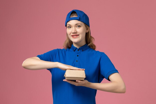 Weiblicher Kurier der Vorderansicht in der blauen Uniform und im Umhang, die kleines Liefernahrungsmittelpaket auf dem rosa Hintergrundlieferuniformuniversitätsarbeitsarbeitsarbeiterjob halten