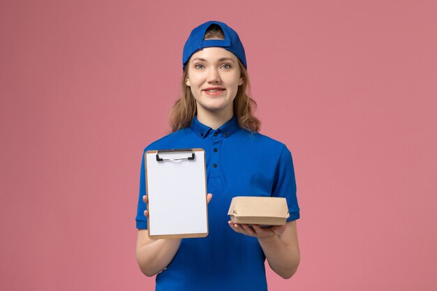 Weiblicher Kurier der Vorderansicht in der blauen Uniform und im Umhang, die kleines Liefernahrungsmittelpaket mit Notizblock auf rosa Wand halten, Lieferservice-Mitarbeiterjobarbeit
