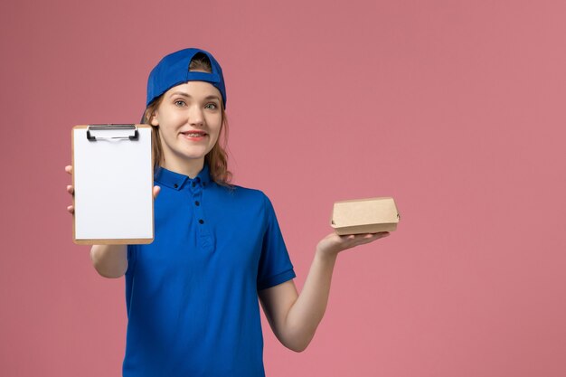 Weiblicher Kurier der Vorderansicht in der blauen Uniform und im Umhang, die kleines Liefernahrungsmittelpaket mit Notizblock auf rosa Wand halten, Zustellungsdienstdienstmitarbeiterarbeit