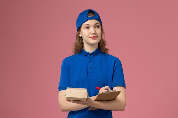 Weiblicher Kurier der Vorderansicht in der blauen Uniform und im Umhang, die kleines Liefernahrungsmittelpaket und Notizblockschreiben auf rosa Wand, Lieferservice-Mitarbeiter halten
