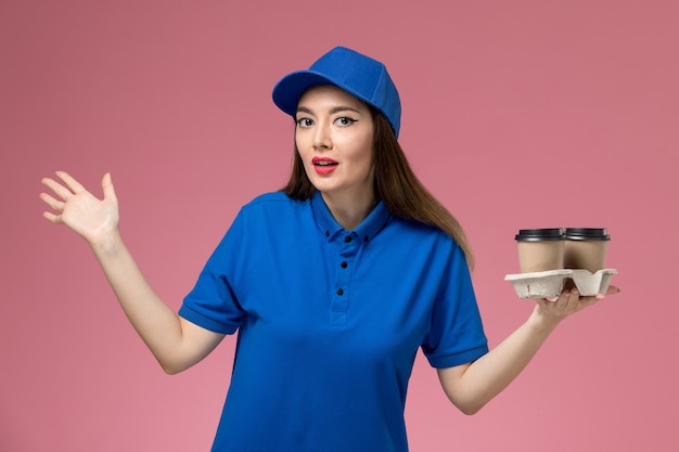 Weiblicher Kurier der Vorderansicht in der blauen Uniform und im Umhang, die Lieferkaffeetassen auf dem rosa Wandjobarbeiter halten