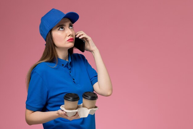 Weiblicher Kurier der Vorderansicht in der blauen Uniform und im Umhang, die Lieferung Kaffeetassen halten, die am Telefon auf rosa Wand sprechen