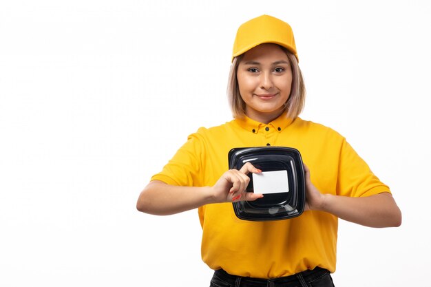 Weiblicher Kurier der Vorderansicht in der gelben Kappe der schwarzen Jeans der gelben Kappe, die Schüsseln mit der weißen Karte des Essens hält, die auf Weiß lächelt