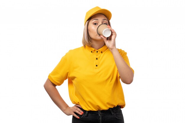 Weiblicher Kurier der Vorderansicht in der gelben Kappe des gelben Hemdes und in den schwarzen Jeans, die Kaffee auf Weiß trinken