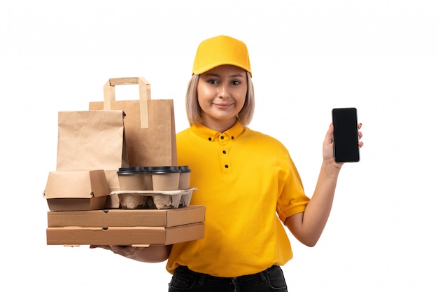 Weiblicher Kurier der Vorderansicht in der gelben Kappe des gelben Hemdes und in den schwarzen Jeans, die Smartphone und Kisten mit dem Essen auf Weiß lächelnd halten