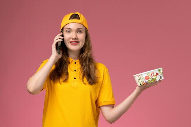 Weiblicher Kurier der Vorderansicht in der gelben Uniform, die Lieferschüssel hält, die durch Telefon auf rosa Wandfirmenservice-Joblieferuniform spricht