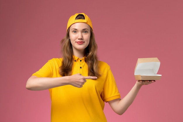 Weiblicher Kurier der Vorderansicht in der gelben Uniform und im Umhang, die kleine Öffnung der Lebensmittelverpackungsöffnung auf dem hellrosa Wanddienst-Lieferuniformjob halten