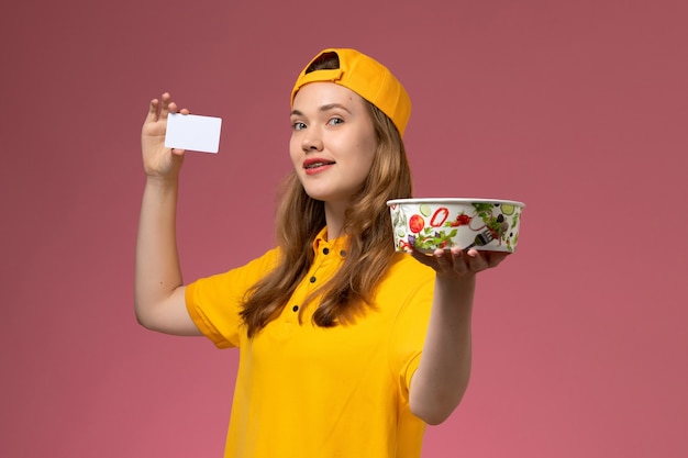 Weiblicher Kurier der Vorderansicht in der gelben Uniform und im Umhang, die Lieferschüssel und Karte auf dem rosa Wanddienstlieferungsuniform-Firmenarbeiterjob halten