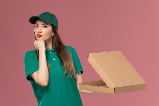 Weiblicher Kurier der Vorderansicht in der grünen Uniform, die Lebensmittel-Lieferbox hält und auf rosa Schreibtischdienstuniform-Lieferauftrag denkt