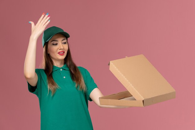 Weiblicher Kurier der Vorderansicht in der grünen Uniform, die Lebensmittellieferungsbo hält, die es auf rosa Wandfirmenserviceuniformlieferung öffnet