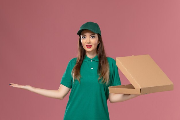 Weiblicher Kurier der Vorderansicht in der grünen Uniform, die Nahrungsmittellieferbox auf der hellrosa Wandfirmenserviceuniformlieferung hält