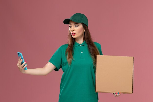 Weiblicher Kurier der Vorderansicht in der grünen Uniform, die Nahrungsmittellieferbox hält und ihr Telefon an der rosa Wandfirmenserviceuniformlieferung verwendet