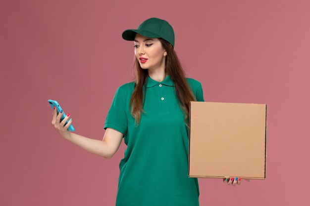 Weiblicher Kurier der Vorderansicht in der grünen Uniform, die Nahrungsmittellieferbox unter Verwendung des Telefons auf der rosa Wandfirmenserviceuniformlieferung hält