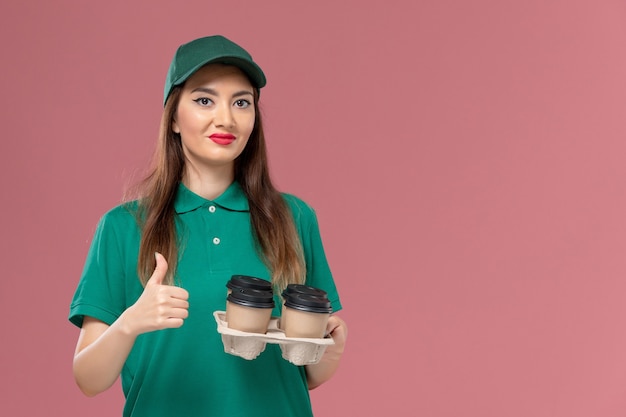 Weiblicher Kurier der Vorderansicht in der grünen Uniform und im Umhang, der die Kaffeetassen der Lieferung auf dem einheitlichen Lieferauftrag der rosa Wanddienstfirma hält