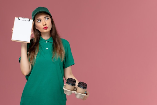 Weiblicher Kurier der Vorderansicht in der grünen Uniform und im Umhang, der Notizblock und Lieferkaffeetassen hält, die auf dem rosa Wanddienstuniform-Lieferjobarbeiter denken