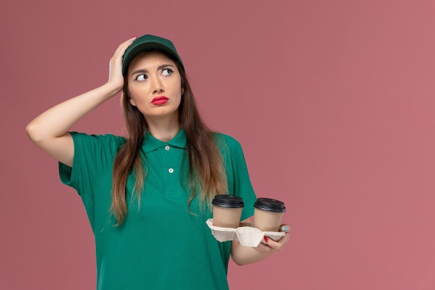 Weiblicher Kurier der Vorderansicht in der grünen Uniform und im Umhang, die Lieferung Kaffeetassen halten, die auf rosa Wandfirmenservicejobuniformlieferung denken