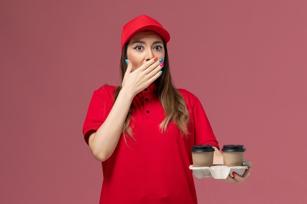 Weiblicher Kurier der Vorderansicht in der roten Uniform, die braune Lieferkaffeetassen auf hellrosa Hintergrunddienstlieferungsuniformjob weiblicher Firma hält