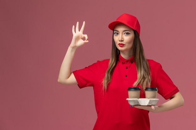 Weiblicher Kurier der Vorderansicht in der roten Uniform, die braune Lieferkaffeetassen hält, die auf rosa Hintergrunddienstjoblieferuniform aufwerfen