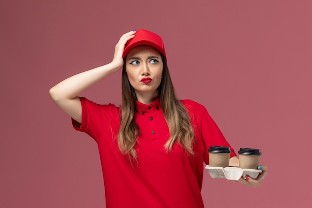 Weiblicher Kurier der Vorderansicht in der roten Uniform, die braune Lieferkaffeetassen hält und an hellrosa Hintergrunddienstlieferungsuniformarbeiterjob weibliches Unternehmen denkt