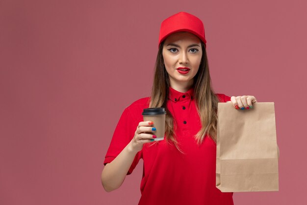 Weiblicher Kurier der Vorderansicht in der roten Uniform, die die Kaffeetasse und das Lebensmittelpaket der Lieferung auf dem rosa Schreibtischdienstlieferuniformjob hält