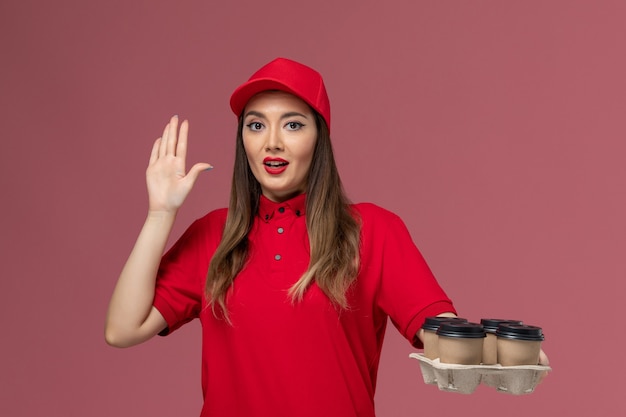 Kostenloses Foto weiblicher kurier der vorderansicht in der roten uniform, die kaffeetassen der lieferung auf der hellrosa hintergrundarbeitsarbeitsdienst-lieferuniform hält