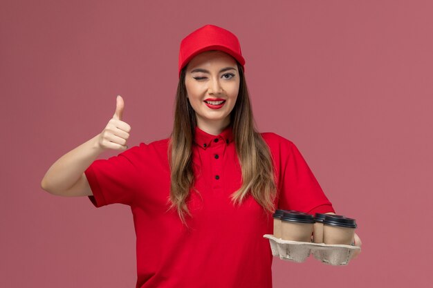 Weiblicher Kurier der Vorderansicht in der roten Uniform, die Lieferkaffeetassen hält, die auf rosa Hintergrundarbeiterjobdienstlieferuniform zwinkert