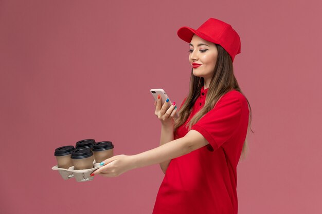 Weiblicher Kurier der Vorderansicht in der roten Uniform, die Lieferkaffeetassen hält, die Foto von ihnen auf rosa Hintergrunddienstlieferungs-Jobuniform machen