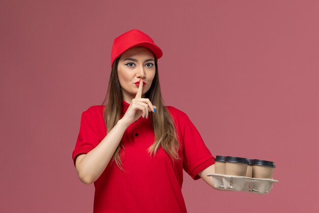 Weiblicher Kurier der Vorderansicht in der roten Uniform, die Lieferkaffeetassen hält, die Stillezeichen auf dem rosa Hintergrunddienstlieferungsuniformjob zeigen