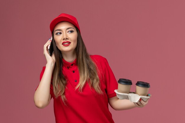 Weiblicher Kurier der Vorderansicht in der roten Uniform, die Lieferkaffeetassen hält und am Telefon auf der rosa Hintergrunddienstlieferuniform spricht
