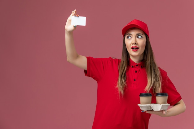 Weiblicher Kurier der Vorderansicht in der roten Uniform, die Lieferkaffeetassen und -karte auf der rosa Hintergrunddienstauftragslieferuniform hält