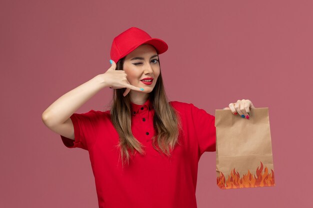 Weiblicher Kurier der Vorderansicht in der roten Uniform, die Papiernahrungsmittelpaket auf der rosa Hintergrundarbeiterservicejobzustellungsuniformfirma hält