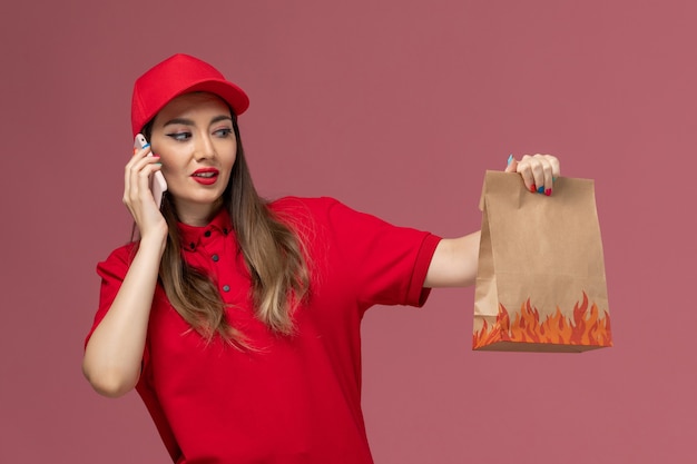 Weiblicher Kurier der Vorderansicht in der roten Uniform, die Telefon und Lebensmittelpaket auf rosa Hintergrunddienstlieferungsuniform-Firmenarbeiter hält