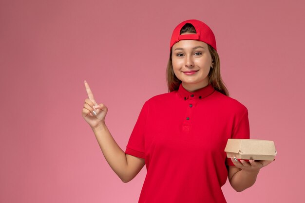 Weiblicher Kurier der Vorderansicht in der roten Uniform und im Umhang, der das Liefernahrungsmittelpaket an der rosa Wand hält, einheitliches Lieferservicejobarbeiterarbeitsmädchen