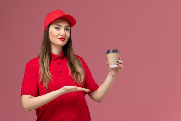 Weiblicher Kurier der Vorderansicht in der roten Uniform und im Umhang, der die Kaffeetasse der Lieferung auf dem hellrosa Hintergrunddienstlieferuniform-Jobarbeiter hält