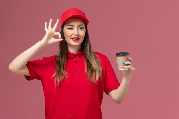 Weiblicher Kurier der Vorderansicht in der roten Uniform und im Umhang, der die Kaffeetasse der Lieferung hält, die auf dem rosa Hintergrunddienstlieferungsuniform-Jobarbeiter zwinkert