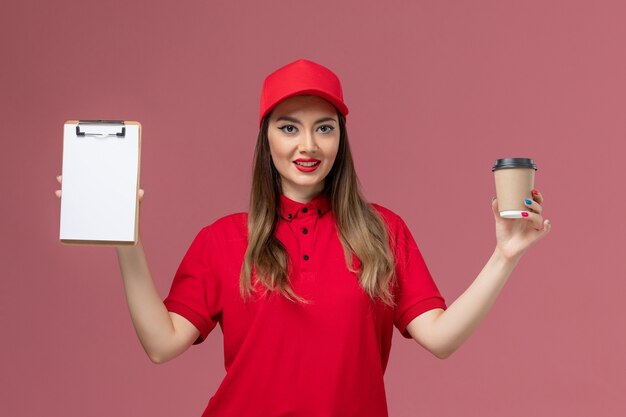 Weiblicher Kurier der Vorderansicht in der roten Uniform und im Umhang, der die Kaffeetasse der Lieferung mit Notizblock auf der rosa Hintergrunddienstlieferungsuniform hält