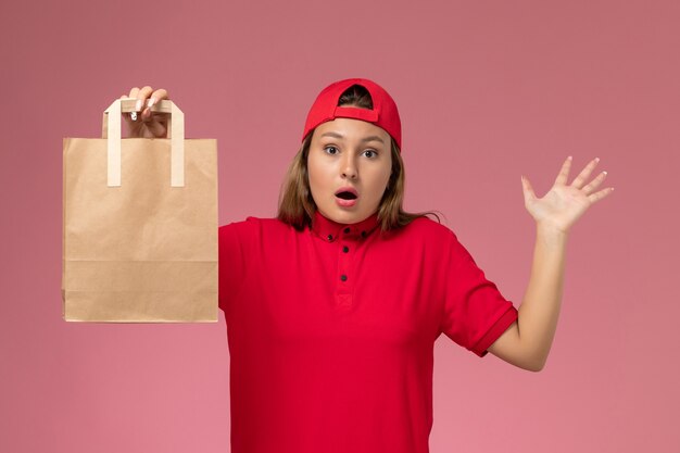 Weiblicher Kurier der Vorderansicht in der roten Uniform und im Umhang, der Lieferpapierpaket an der rosa Wand hält, einheitliche Lieferservicearbeit