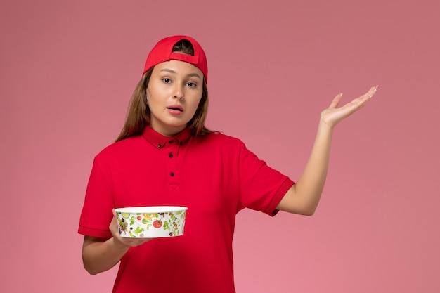 Weiblicher Kurier der Vorderansicht in der roten Uniform und im Umhang, der Lieferschüssel an der rosa Wand hält, einheitlicher Lieferservicejob