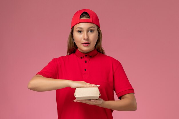 Weiblicher Kurier der Vorderansicht in der roten Uniform und im Umhang, der Lieferung-Nahrungsmittelpaket an der rosa Wand hält, Arbeitsmädchen des Uniform-Lieferserviceunternehmens
