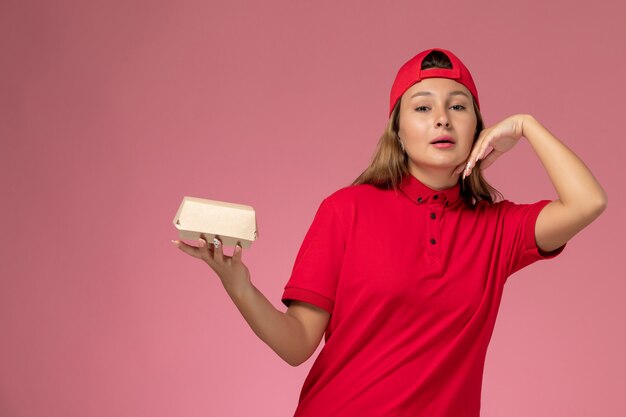 Weiblicher Kurier der Vorderansicht in der roten Uniform und im Umhang, der Lieferung-Nahrungsmittelpaket hält, das auf rosa Wand, Arbeiteruniform-Lieferservice-Firmenjob aufwirft