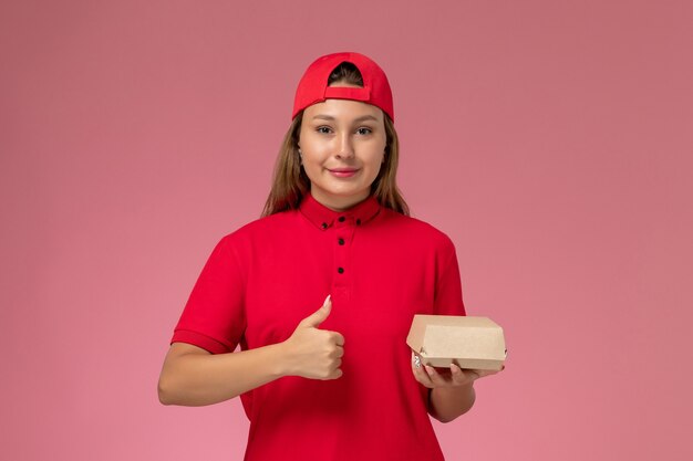 Weiblicher Kurier der Vorderansicht in der roten Uniform und im Umhang, der Lieferung-Nahrungsmittelpaket hält und auf der rosa Wand lächelt, einheitlicher Lieferservice-Firmenjob