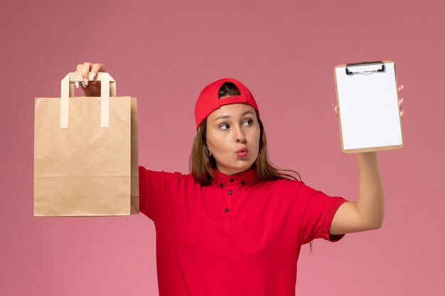 Weiblicher Kurier der Vorderansicht in der roten Uniform und im Umhang, der Lieferung-Nahrungsmittelpaket mit Notizblock auf hellrosa Wand hält, einheitlicher Zustellungsjobdienst