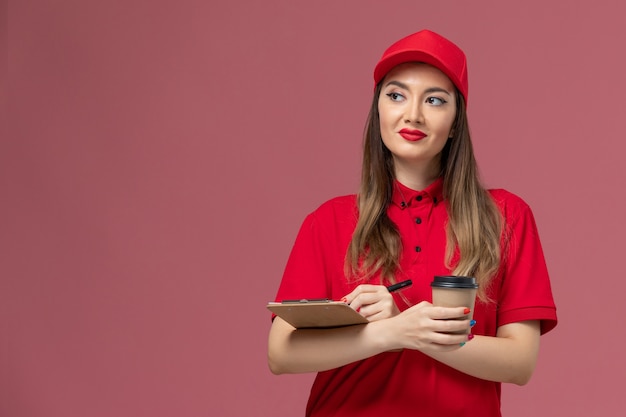 Weiblicher Kurier der Vorderansicht in der roten Uniform und im Umhang, die die Kaffeetasse der Lieferung mit Notizblock und Stift auf hellrosa Hintergrunddienstlieferuniform halten