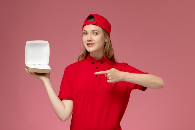 Weiblicher Kurier der Vorderansicht in der roten Uniform und im Umhang, die kleines leeres Liefernahrungsmittelpaket an der rosa Wand halten, Lieferserviceunternehmensuniformjob