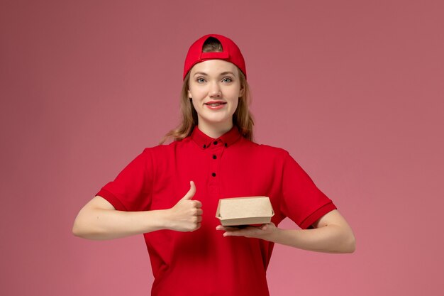 Weiblicher Kurier der Vorderansicht in der roten Uniform und im Umhang, die kleines Liefernahrungsmittelpaket an der rosa Wand halten, Lieferserviceuniformuniformarbeit