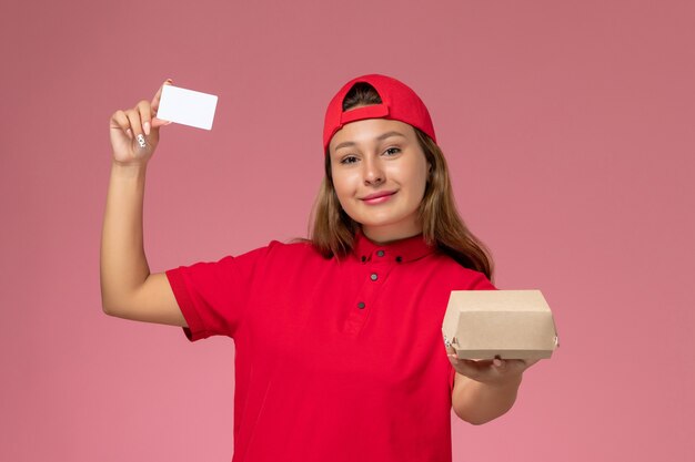 Weiblicher Kurier der Vorderansicht in der roten Uniform und im Umhang, die kleines Liefernahrungsmittelpaket und Karte auf rosa Wand halten, einheitlicher Lieferservicearbeiter