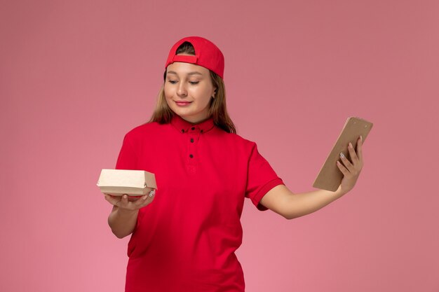 Weiblicher Kurier der Vorderansicht in der roten Uniform und im Umhang, die kleines Liefernahrungsmittelpaket und Notizblock auf rosa Wand halten, einheitliche Lieferarbeiterservicefirma