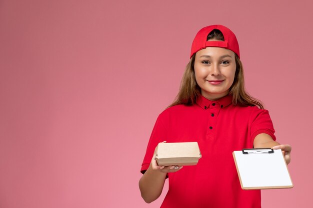 Weiblicher Kurier der Vorderansicht in der roten Uniform und im Umhang, die kleines Liefernahrungsmittelpaket und Notizblock lächelnd auf der rosa Wand, Uniformarbeiter-Lieferservice halten